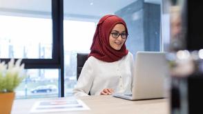A woman sits at a desk, smiling at her laptop