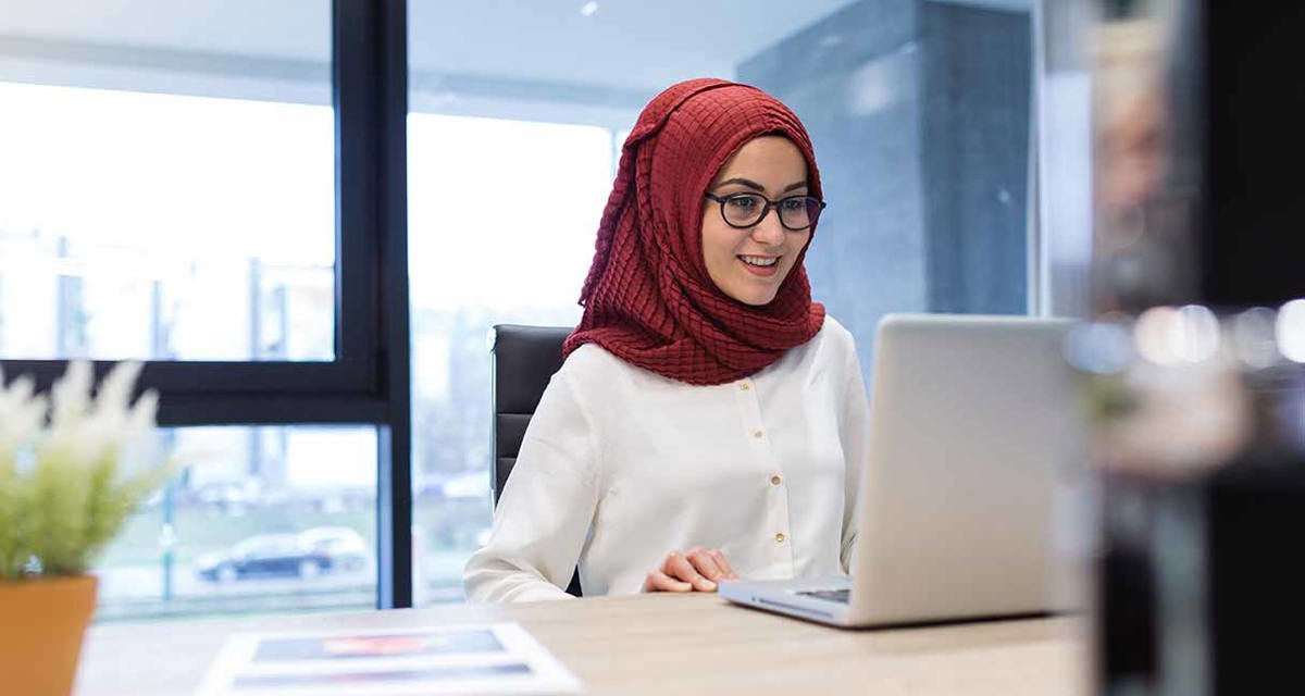 A woman sits at a desk, smiling at her laptop